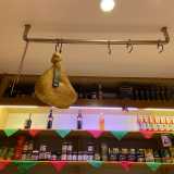 Sullo sfondo si stagliano mensole di vari generi alimentari, mentre da una trave sul soffitto pende una coscia di prosciutto crudo
